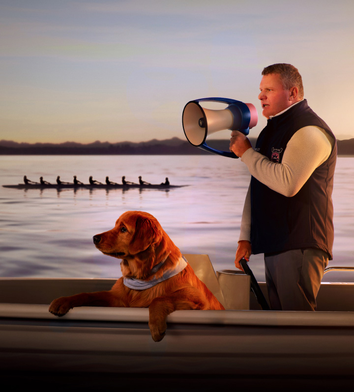 Guy Monseair, junto a un lago con un perro, entrena a un equipo de remo con un megáfono en la mano