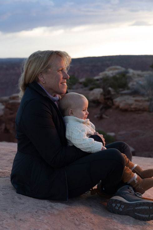 Barbara sitting with baby Jack, who is wearing a warm beige sweater, in the mountains at sunset.