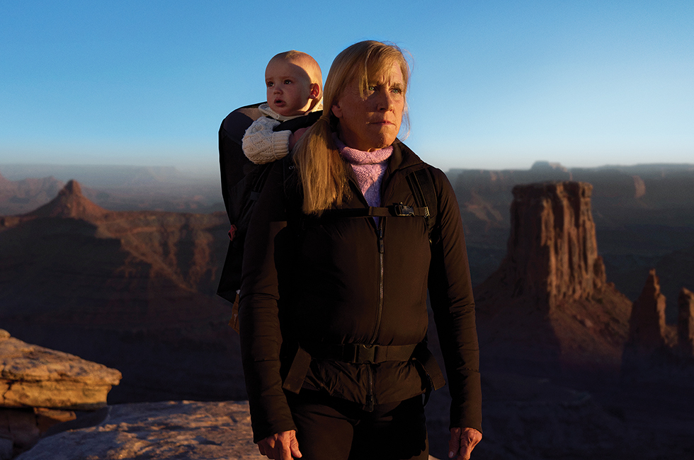 Barbara with baby Jack in a backpack on the mountaintop. Barbara stares into the distance resolutely.