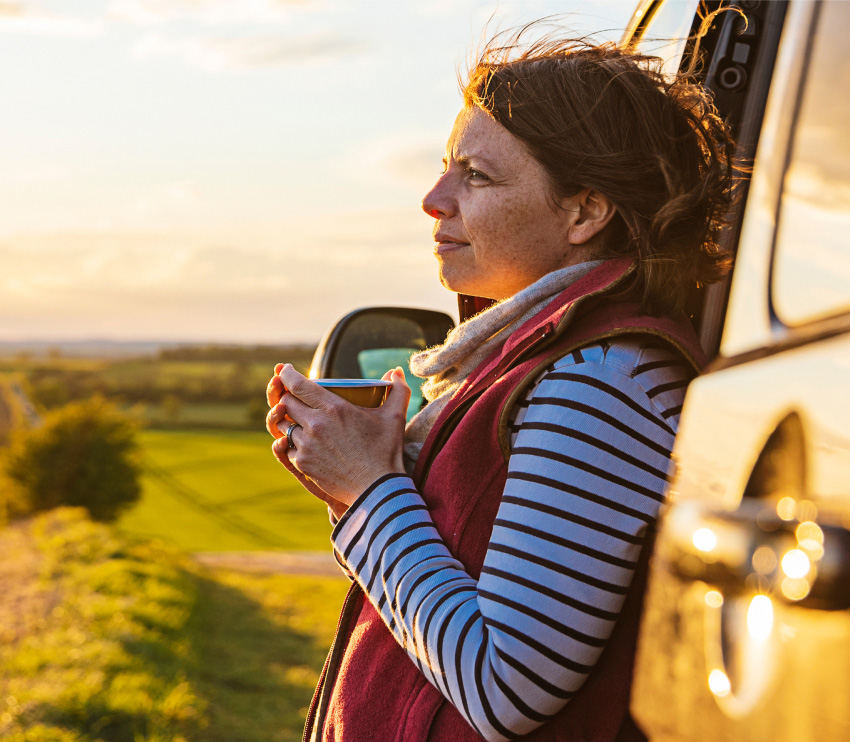 Woman with cup of coffee leaning against a pickup truck in a pastoral setting